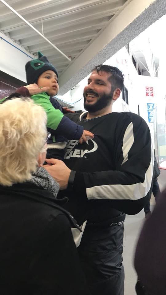 Image of white male in a black and white hockey uniform holding a young child wearing a green hat.
